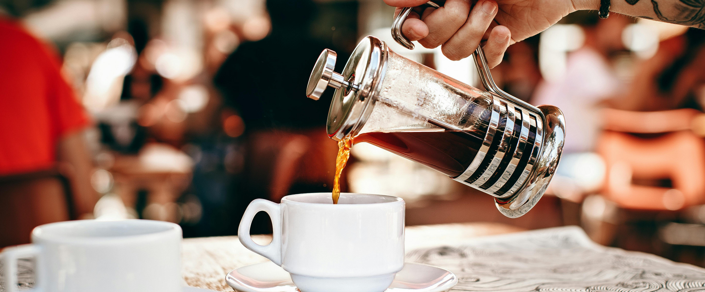 Hand pouring coffee from French press into a cup on a table.