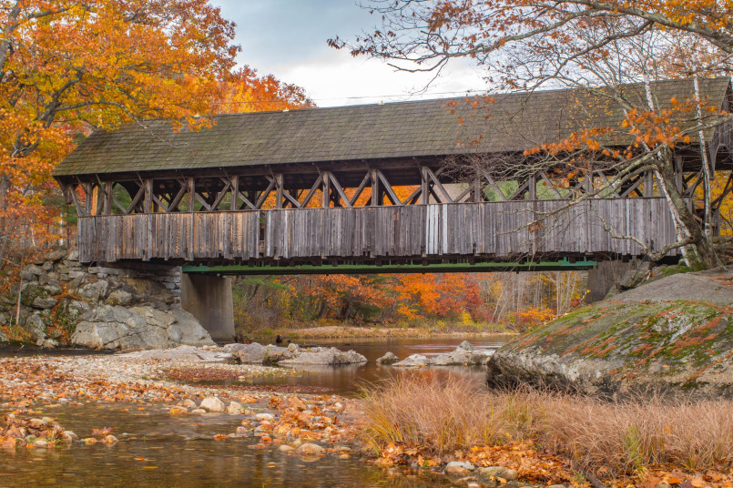 White covered bridge over a rocky river with autumn foliage in Maine