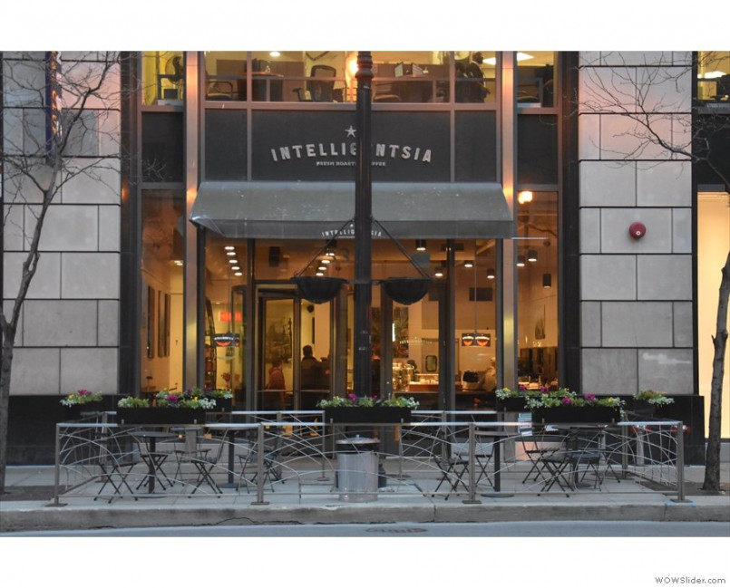 A street view of a coffee shop with outdoor seating. The shop has large glass windows and a black awning with the name 'Intelligentsia' visible. There are tables and chairs set up outside with flower planters around.