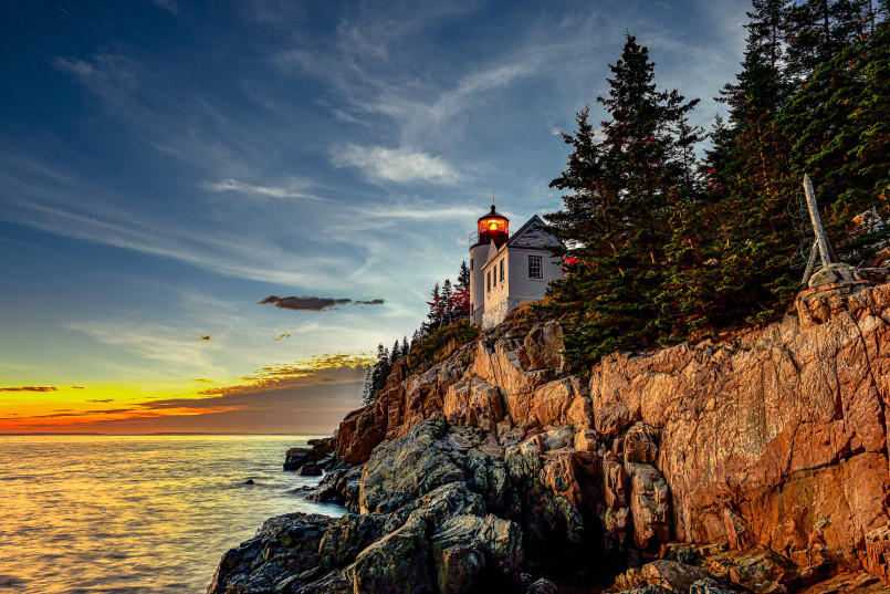 Bass Harbor Head Lighthouse Acadia National Park Maine sunset Small white lighthouse with red roof among evergreen trees at rocky shoreline