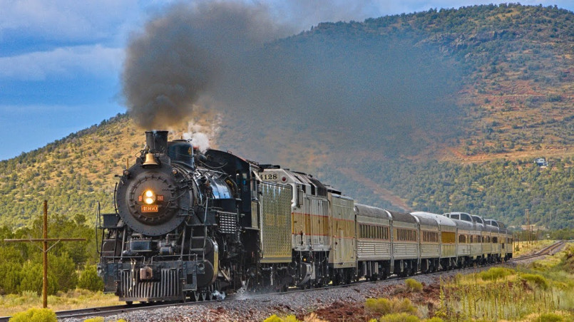Historic Grand Canyon Railway train with red and silver cars traveling through the Arizona high desert with mesas in the background